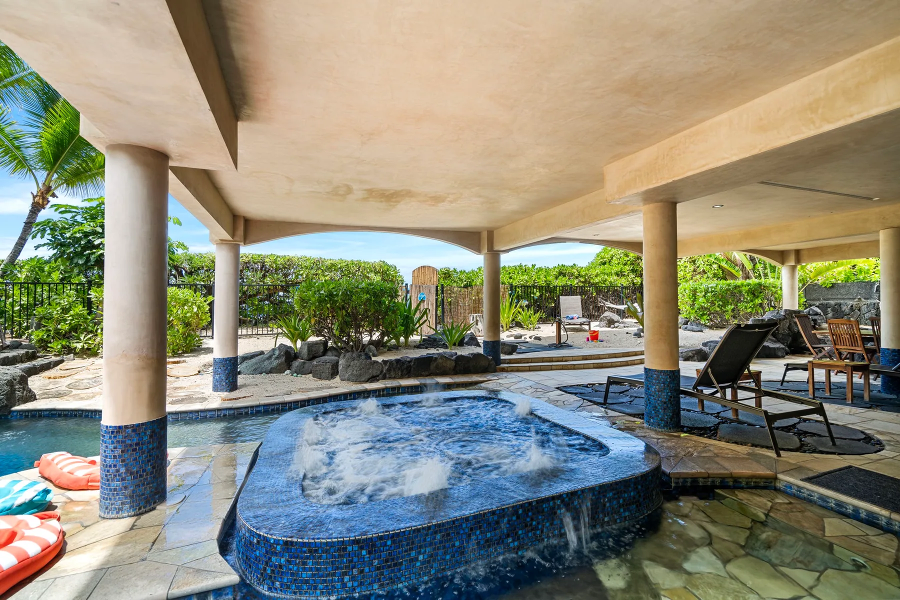 Outdoor pool beneath a covered patio, with lounge seating and tropical greenery.