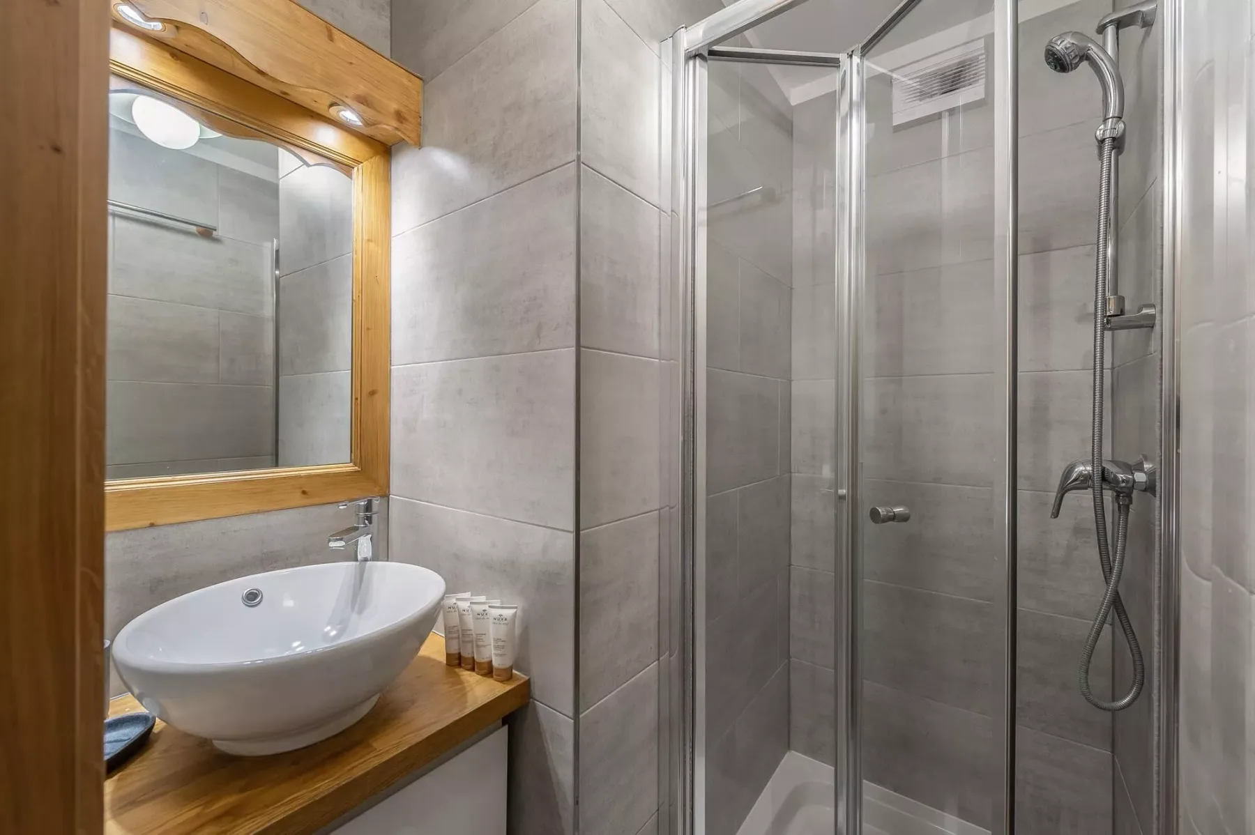 Un baño alicatado con ducha de vidrio y un lavabo bajo un espejo con marco de madera.