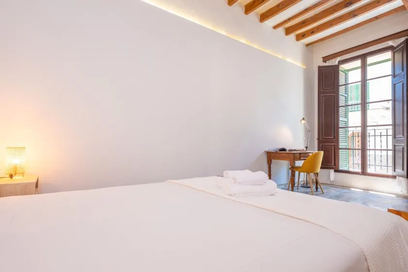 Un dormitorio luminoso tiene una cama grande y un escritorio junto a ventanas altas.