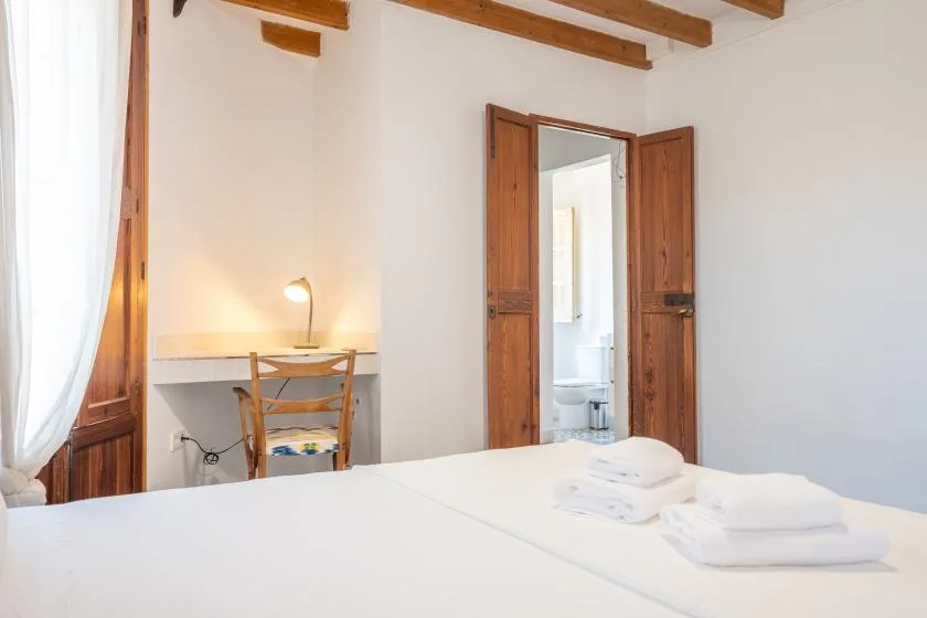 Un dormitorio luminoso con escritorio, vigas de madera en el techo y una puerta abierta al baño.