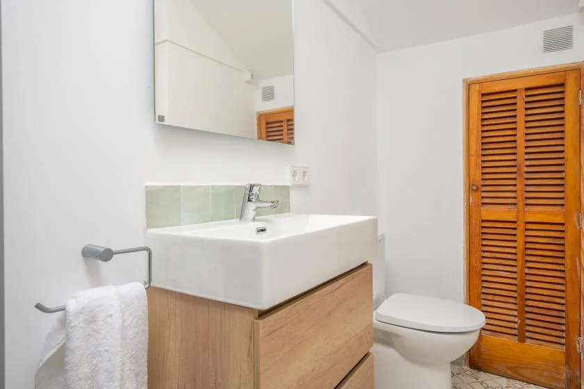 Baño con lavabo blanco, mueble de madera e inodoro junto a una puerta de celosía.