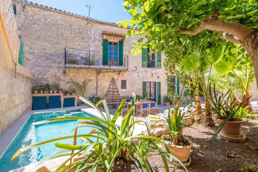Una piscina en un patio junto a un edificio de piedra, a la sombra de árboles y rodeada de plantas.