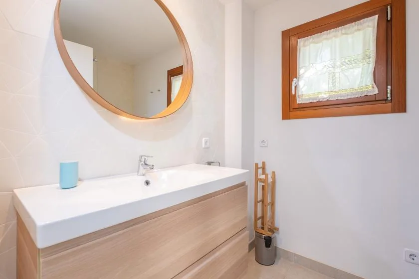 Un lavabo de baño está bajo un espejo redondo, con una ventana pequeña cerca.