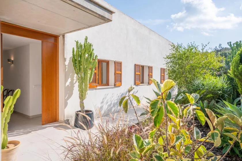 Una pared exterior blanca con ventanas de madera da a un jardín con plantas frondosas.