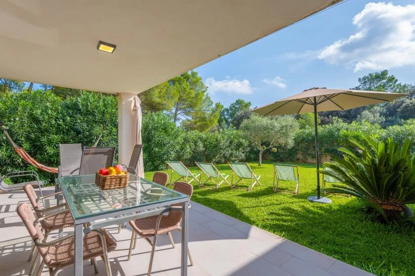 Un patio cubierto con una mesa de comedor de vidrio da a un jardín verde con sillas de jardín.