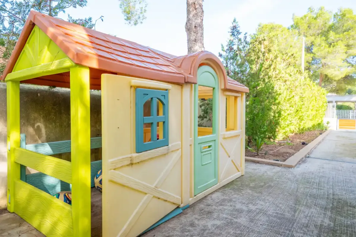 Una casita de juegos infantil de colores está junto a un camino pavimentado en un jardín.