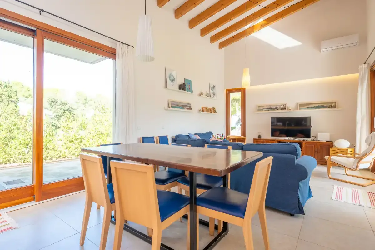 Una luminosa sala de estar y comedor tiene asientos azules, una mesa de madera y grandes puertas de vidrio.