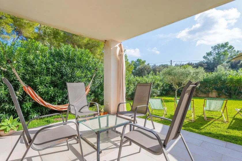Un patio cubierto con asientos, una mesa de cristal y una hamaca da a un jardín verde.