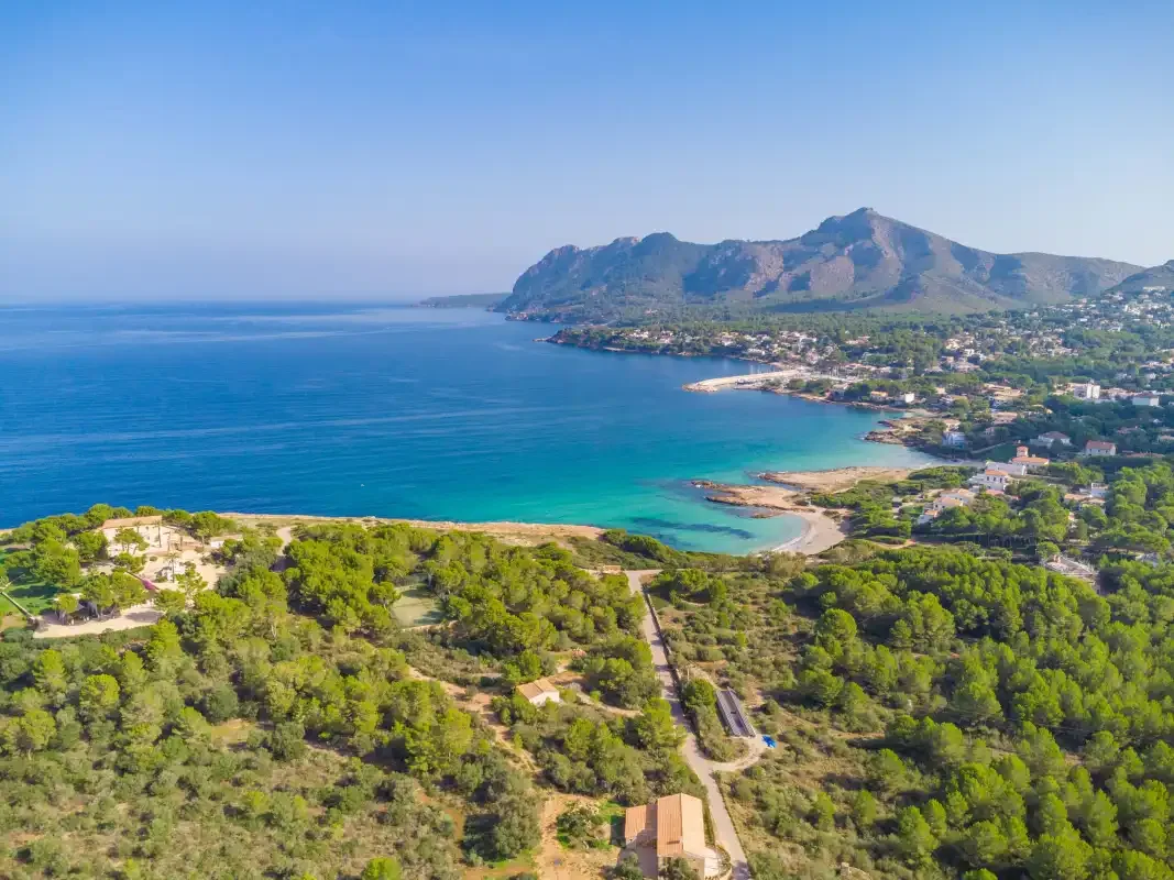 Vista aérea de una bahía costera, con agua turquesa, colinas verdes y una montaña al fondo.