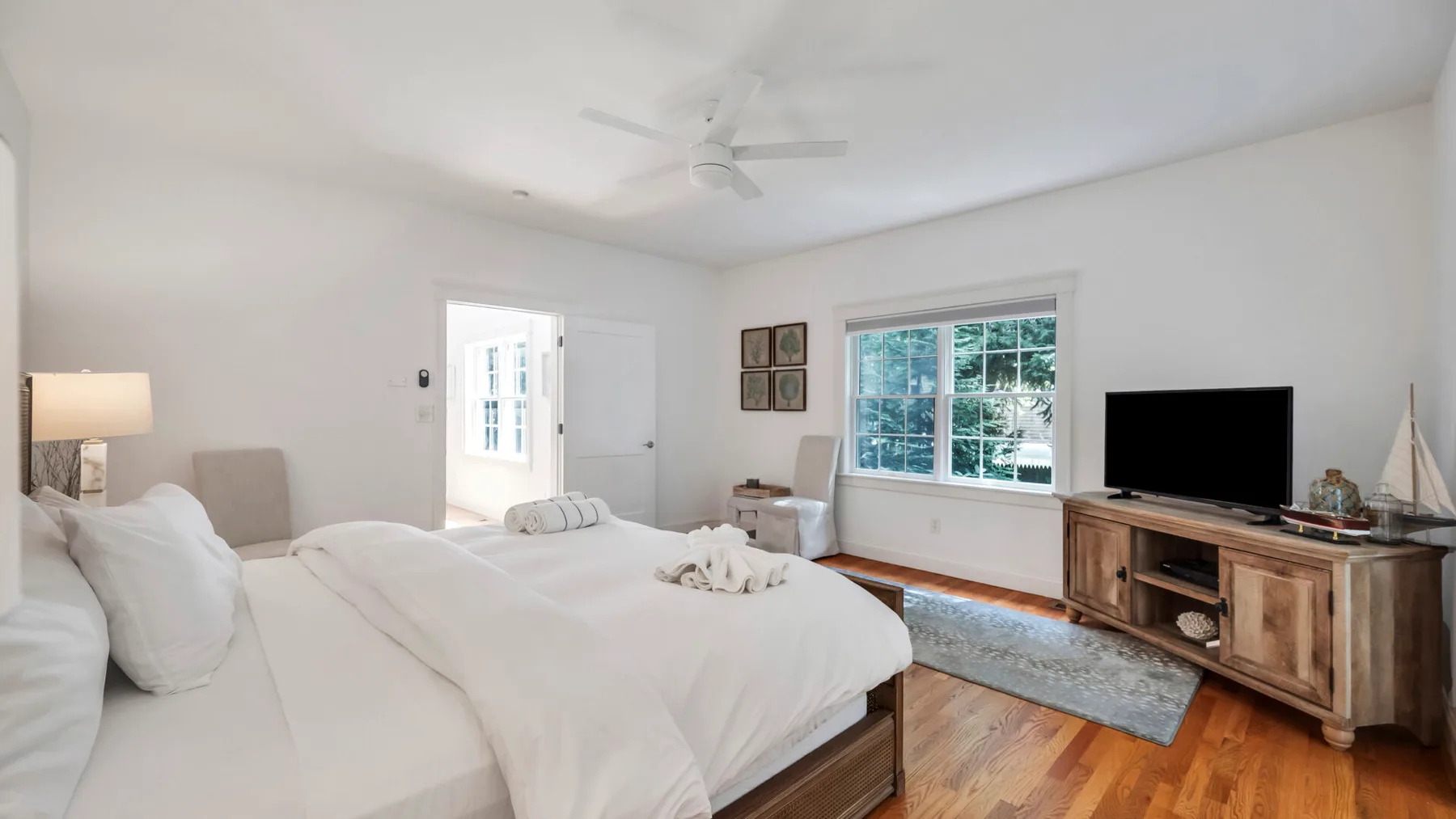 A bright bedroom with white bedding, a large window, and a television on a wooden cabinet.