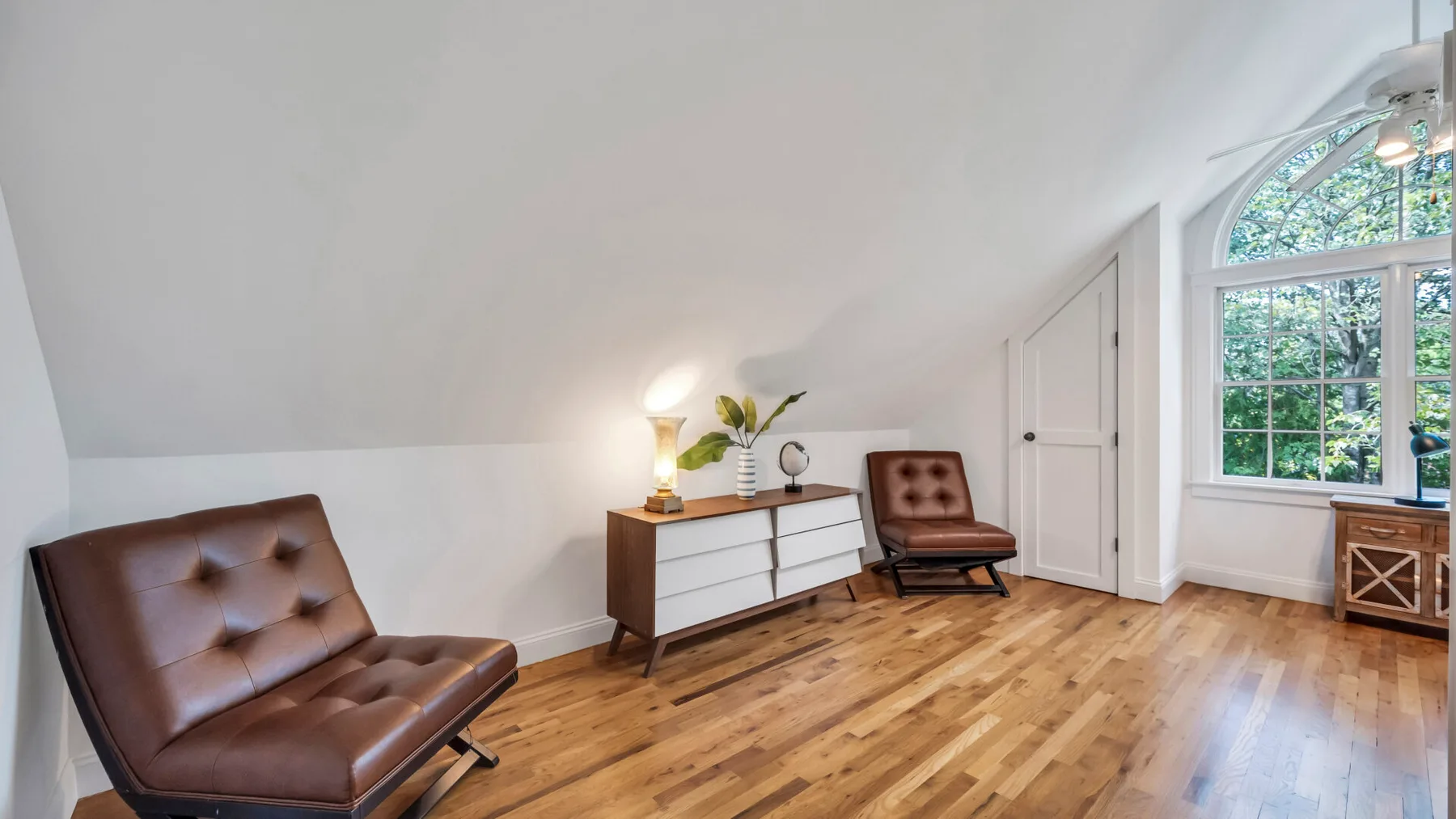 A bright sitting room with brown seating, a white dresser, and a large arched window.