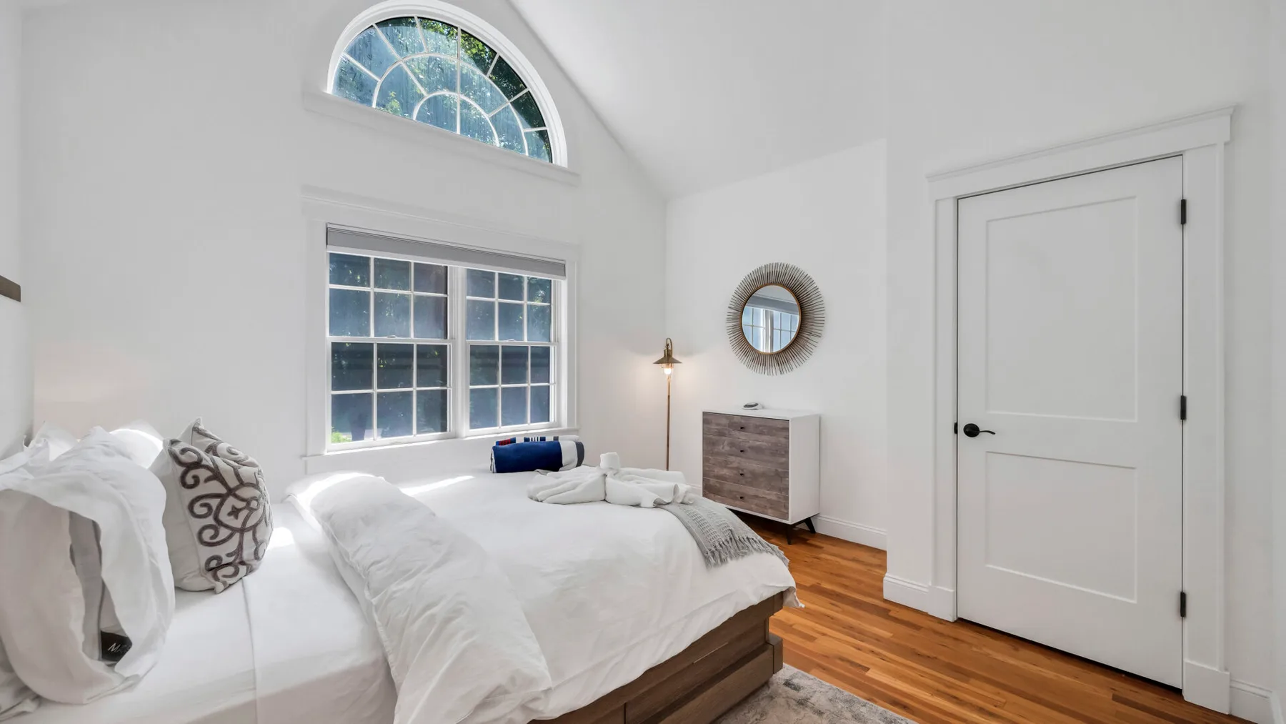 A bright bedroom with white bedding, a large window, and wood flooring.