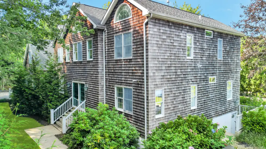 A two-story house with shingled siding stands among trees and shrubs.