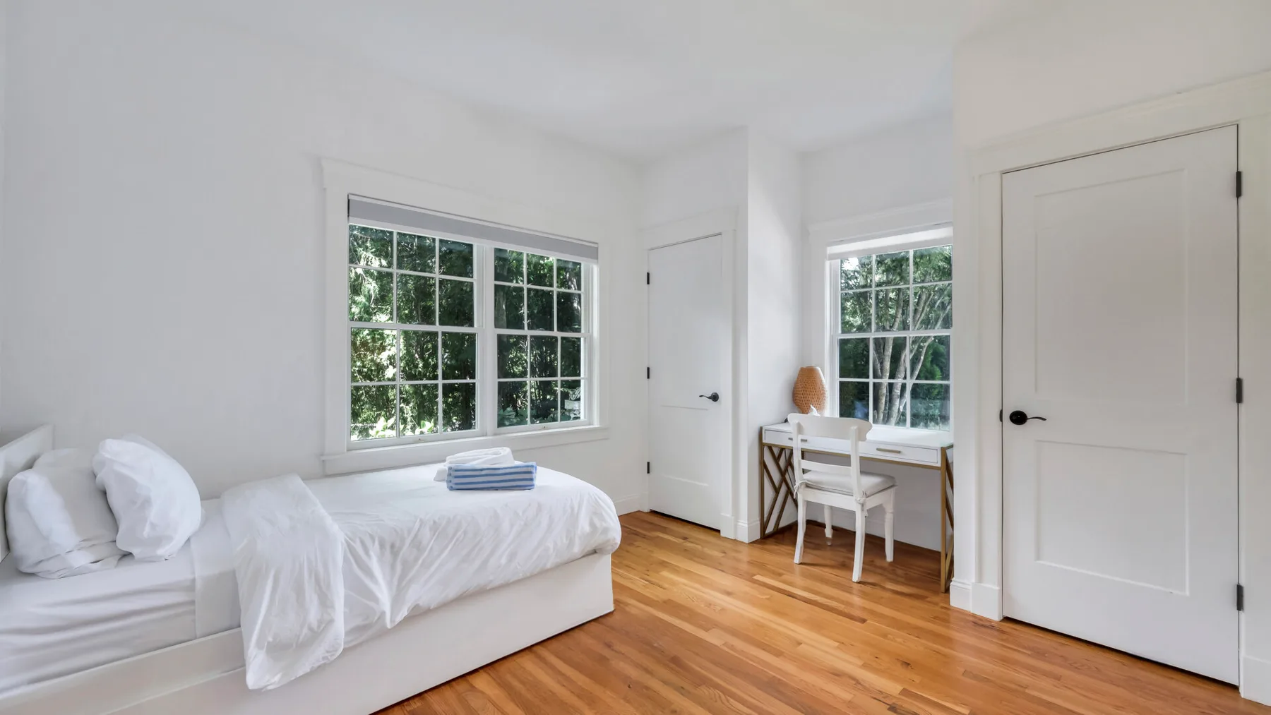 A bright bedroom with a bed, desk, and windows overlooking greenery.