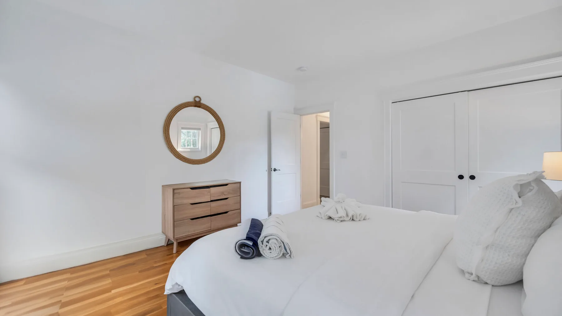 A bright bedroom with a neatly made bed and a wooden dresser beneath a round mirror.