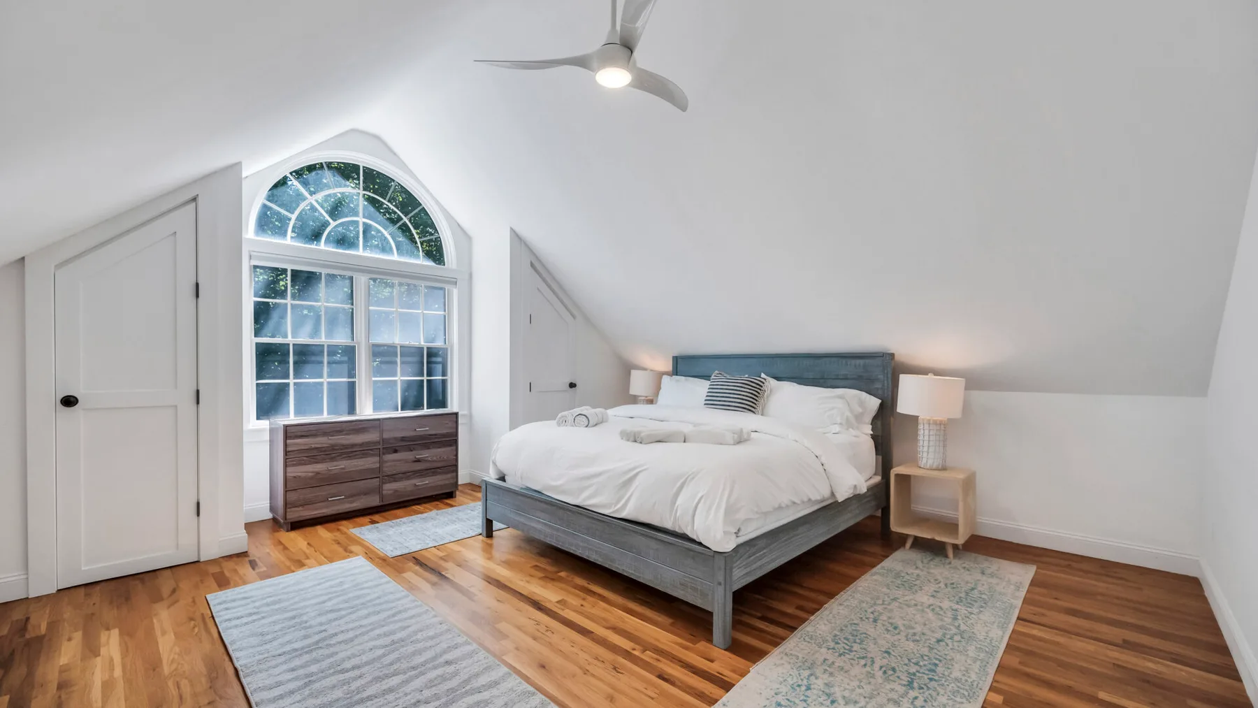 A bright bedroom with a bed beneath a sloped ceiling and an arched window.