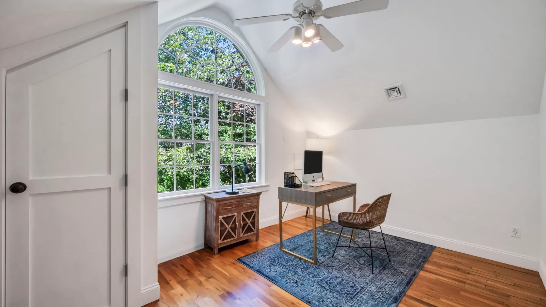 A bright room with an arched window, desk, and seating on a blue rug.