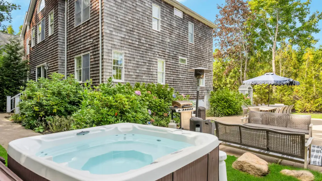 A hot tub and patio seating sit beside a shingled house surrounded by greenery.