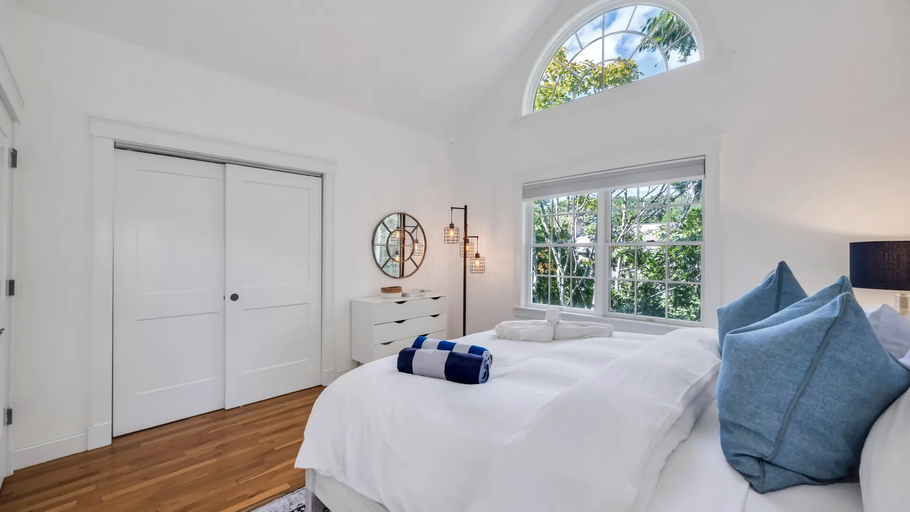 A bright bedroom with white bedding, blue pillows, and large windows.