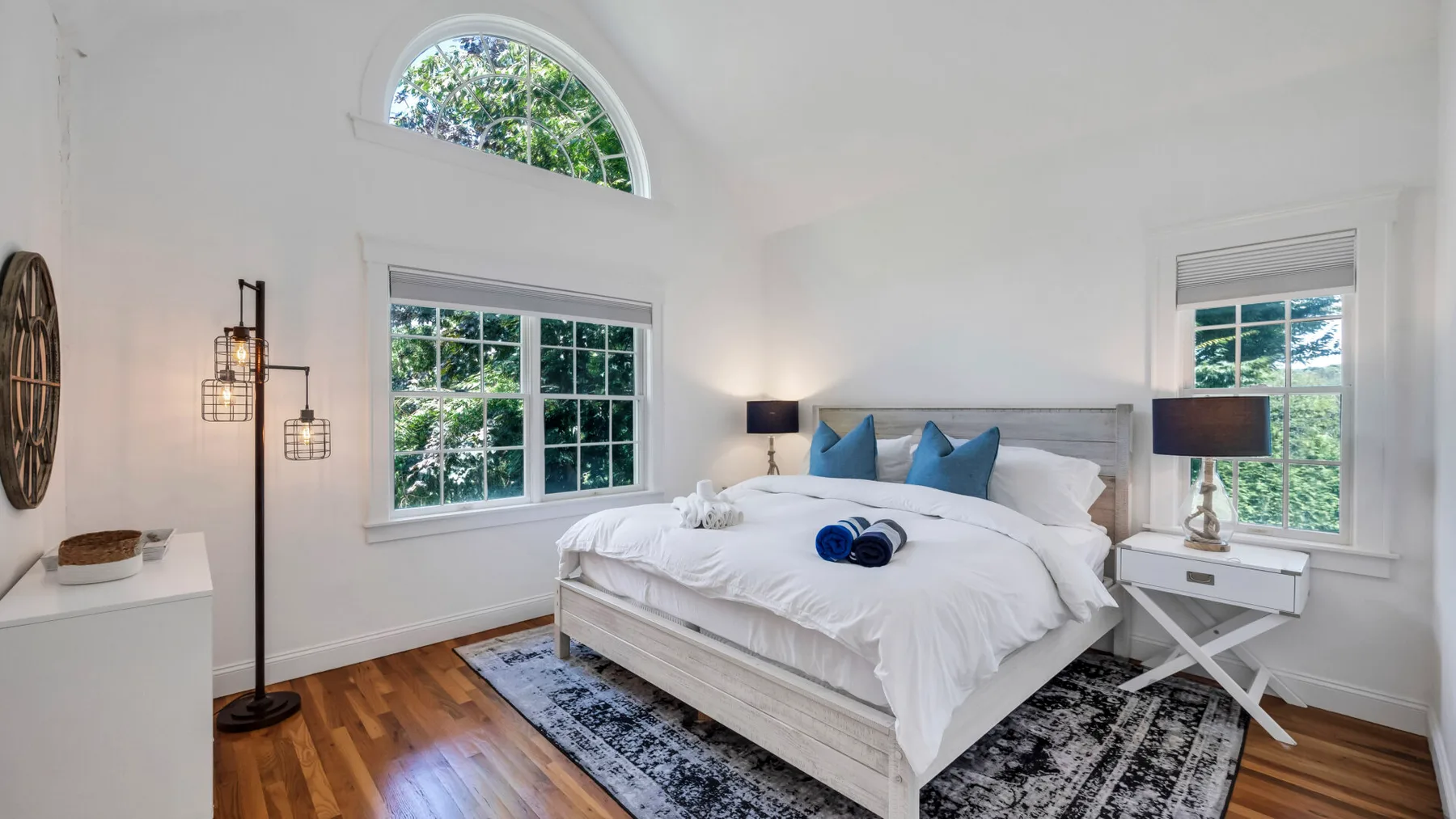 A bright bedroom with a large bed, blue pillows, and windows overlooking greenery.