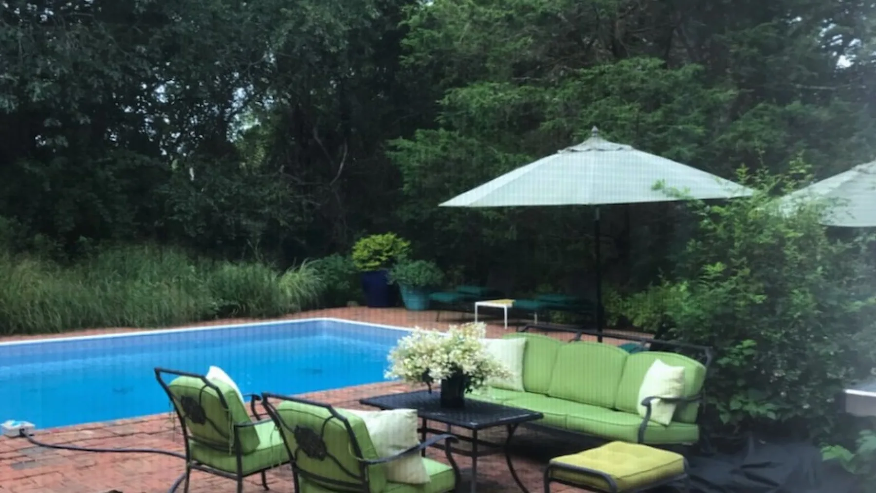 Una piscina al aire libre junto a asientos verdes y una mesa, rodeada de árboles.