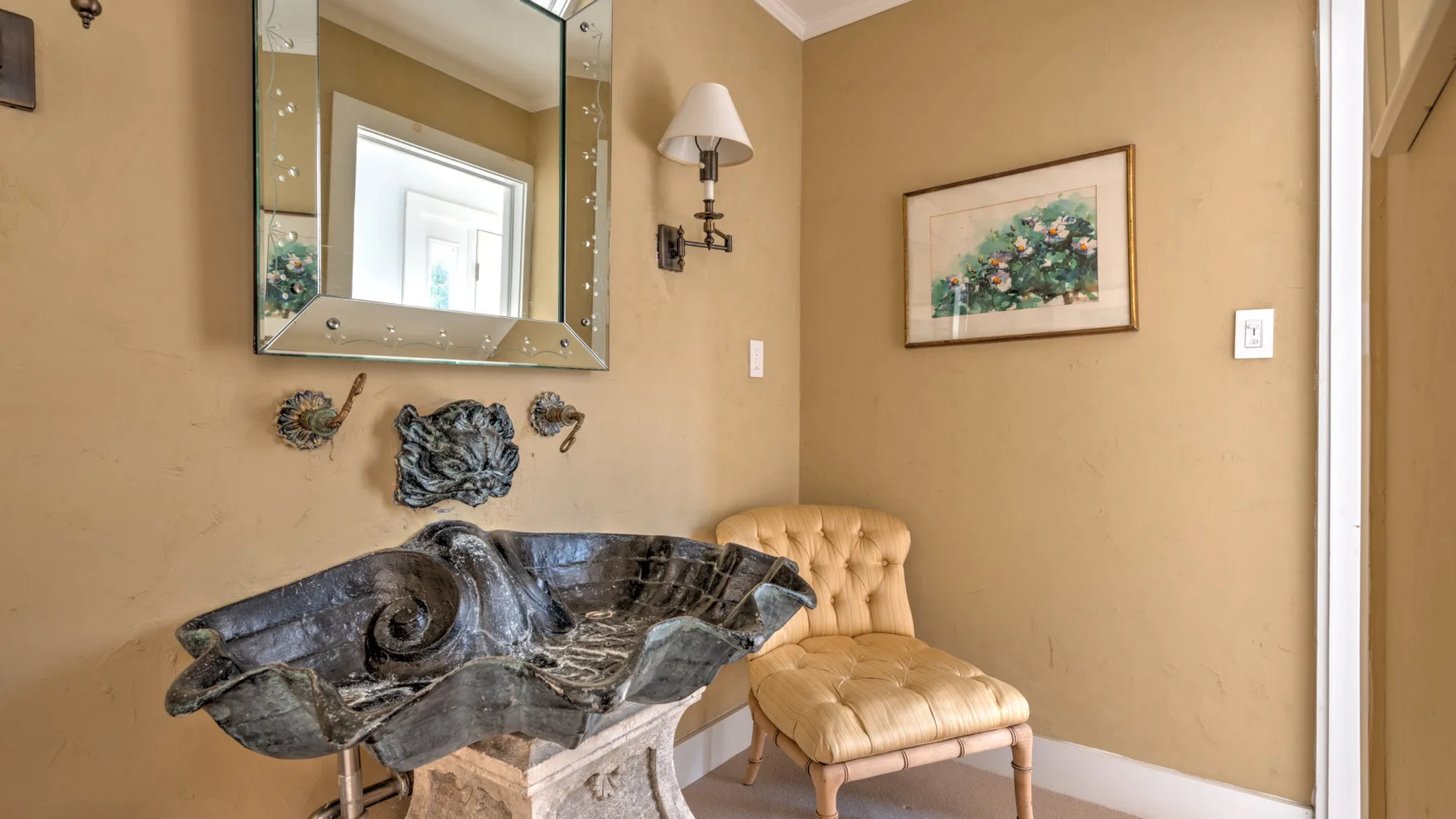 Una habitación beige presenta un lavabo oscuro ornamentado bajo un espejo grande y una silla acolchada cercana.