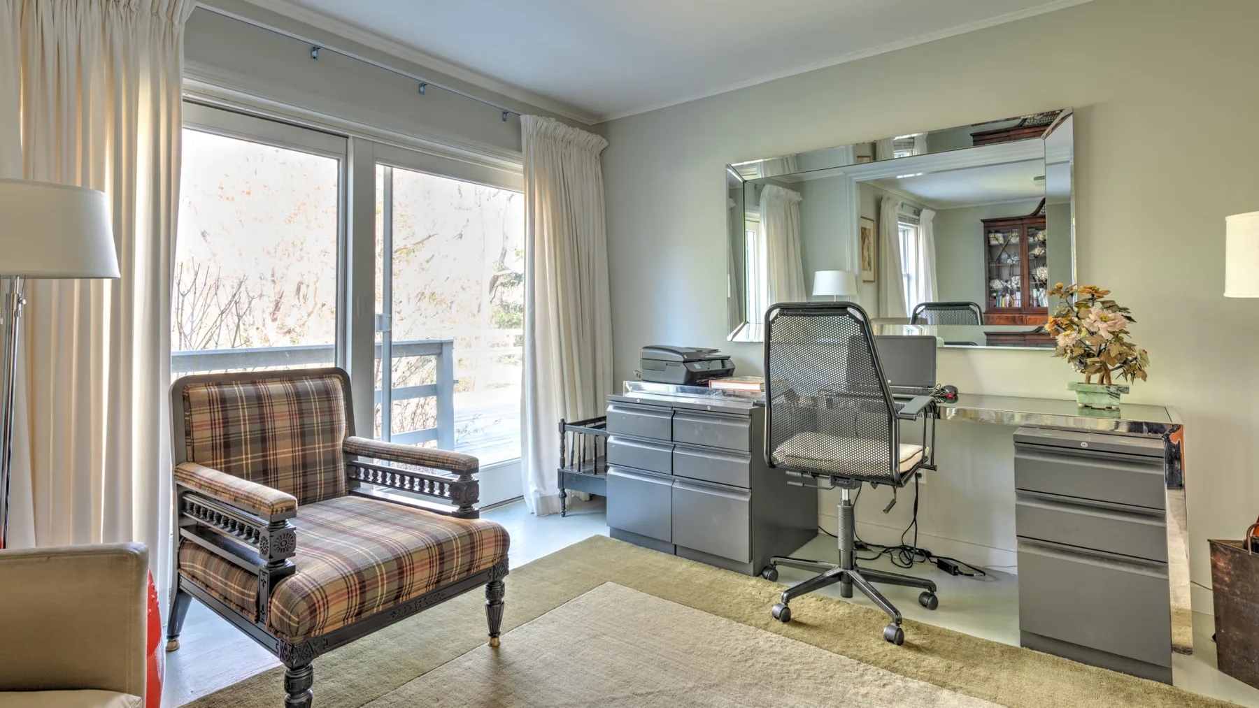 Una habitación luminosa con escritorio, silla de oficina y asiento a cuadros junto a grandes ventanas.