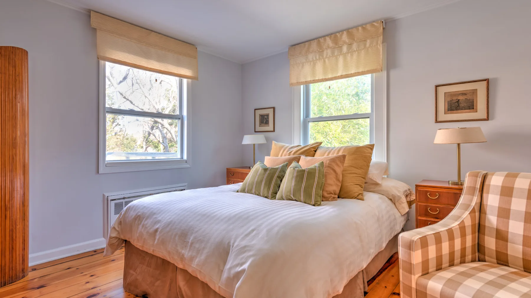 Un dormitorio con una cama tendida, dos ventanas y un sillón a cuadros.