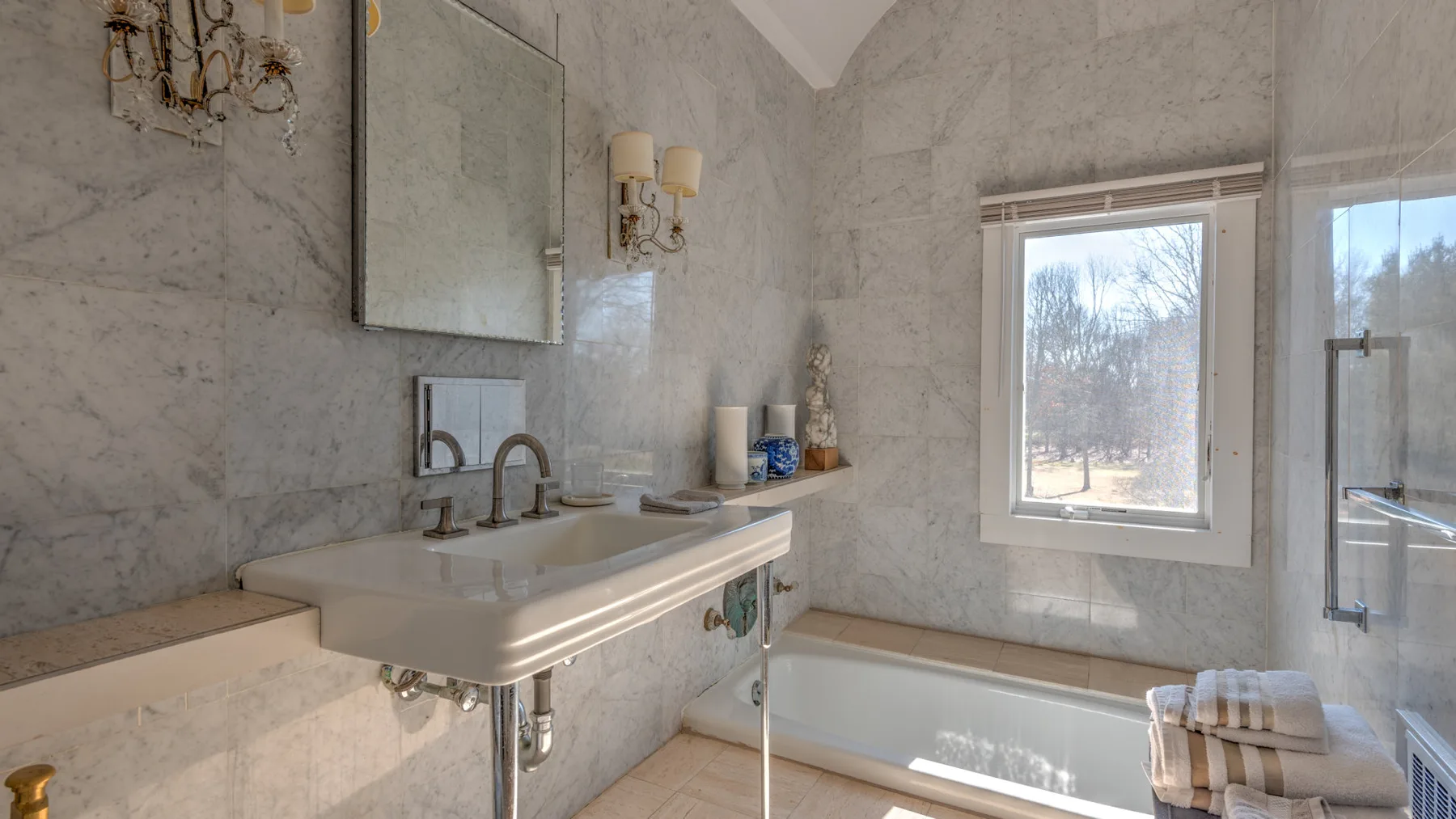 Un baño de mármol con lavabo de pared, bañera y ventanas grandes.