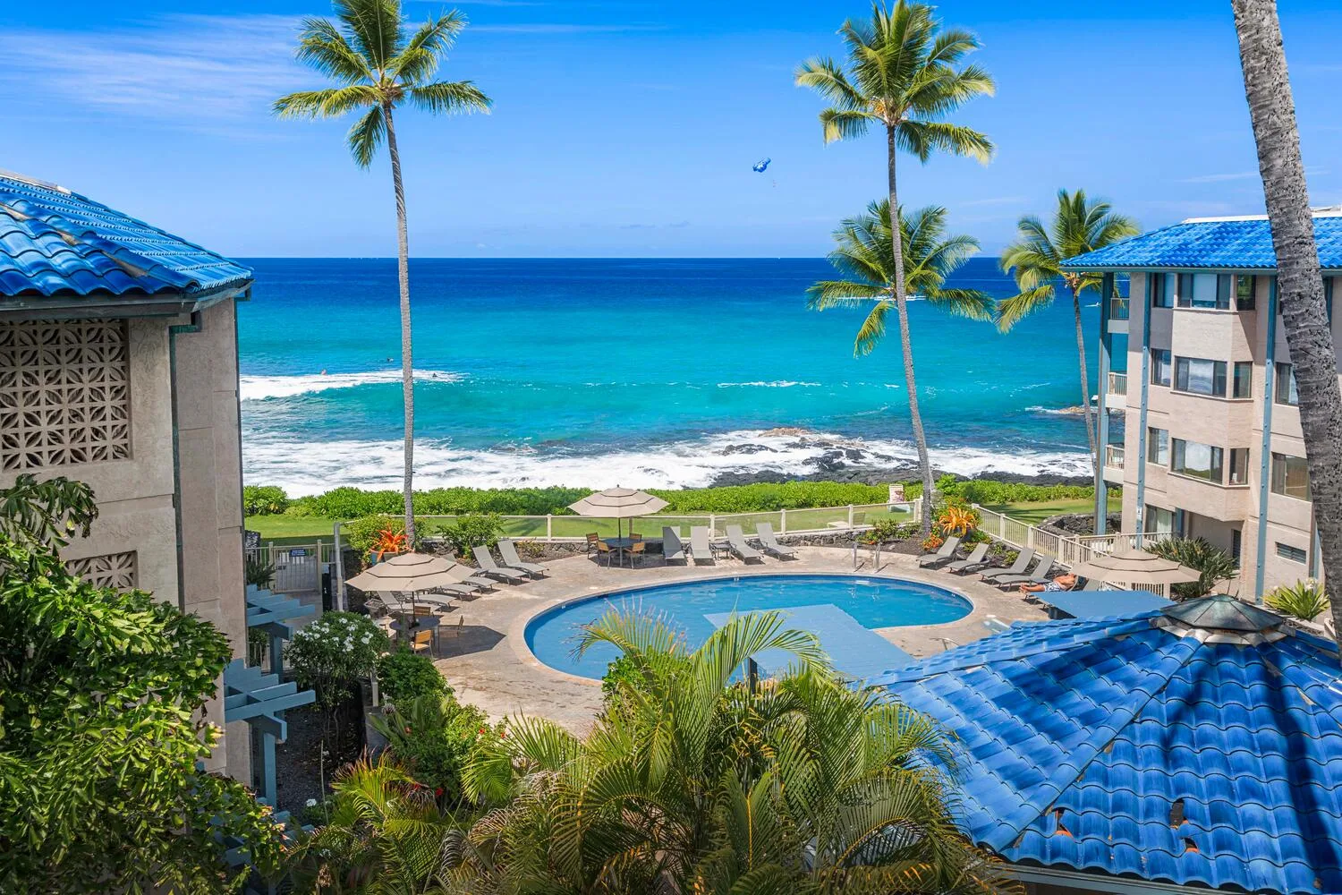 Escena tropical frente al mar con una piscina curva, palmeras y edificios con techos azules.