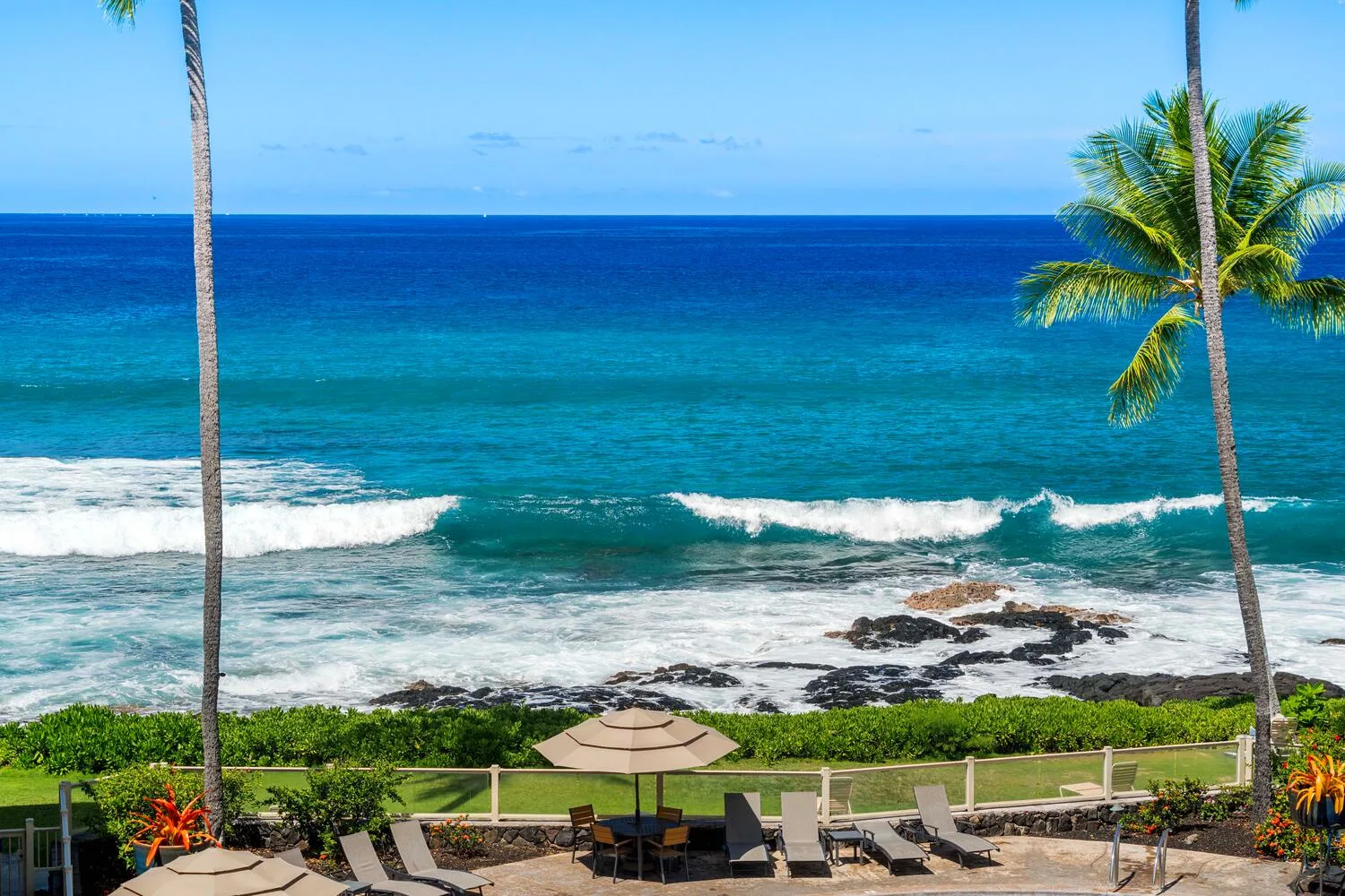 Una vista costera muestra olas turquesas, palmeras y asientos junto a la orilla.