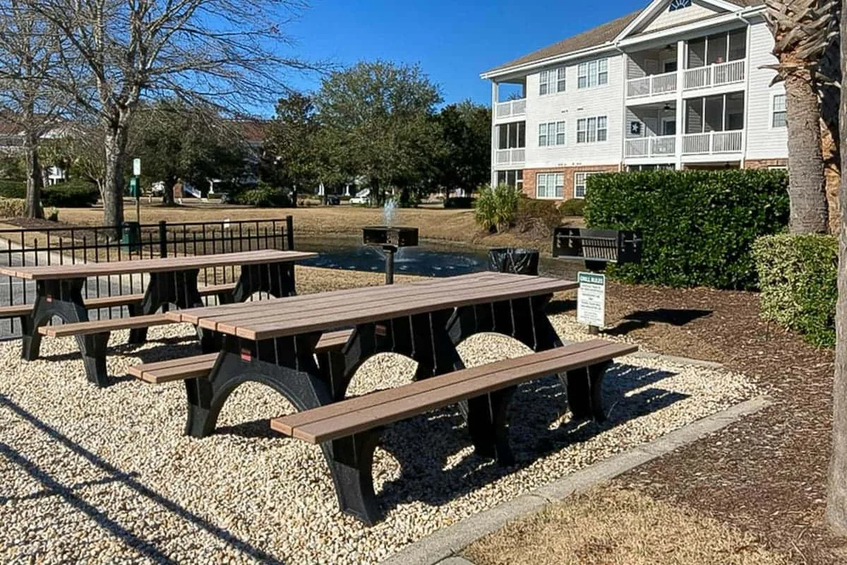Mesas de picnic están en una zona de grava junto a un edificio residencial ajardinado.
