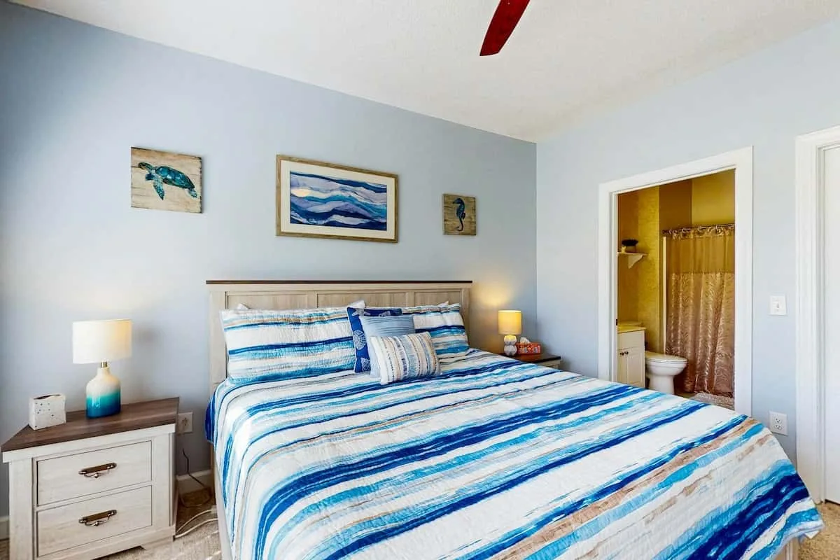 Un dormitorio con una cama cubierta con ropa de cama a rayas azules y blancas junto a una puerta abierta al baño.
