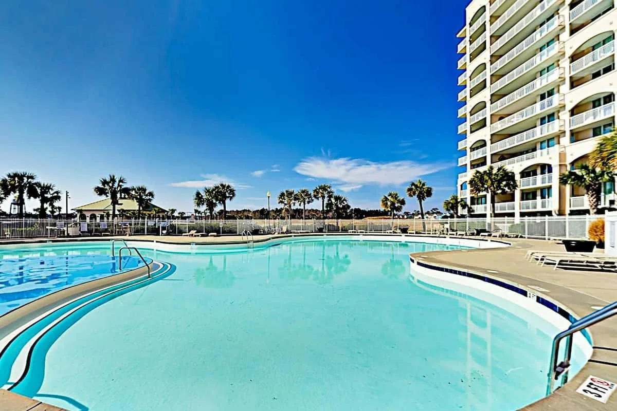 A large outdoor swimming pool sits beside a high-rise building under a clear blue sky.