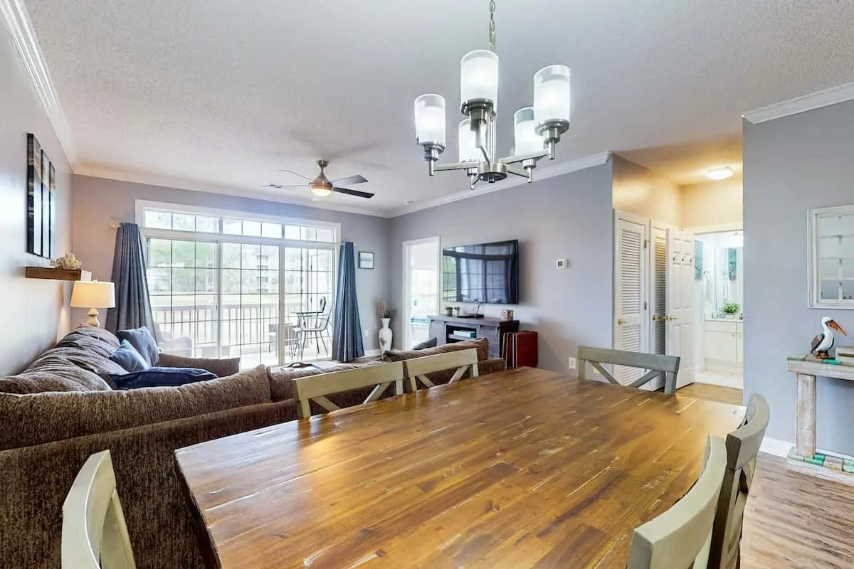Una sala de estar y comedor con una mesa de madera y asientos junto a grandes ventanas.