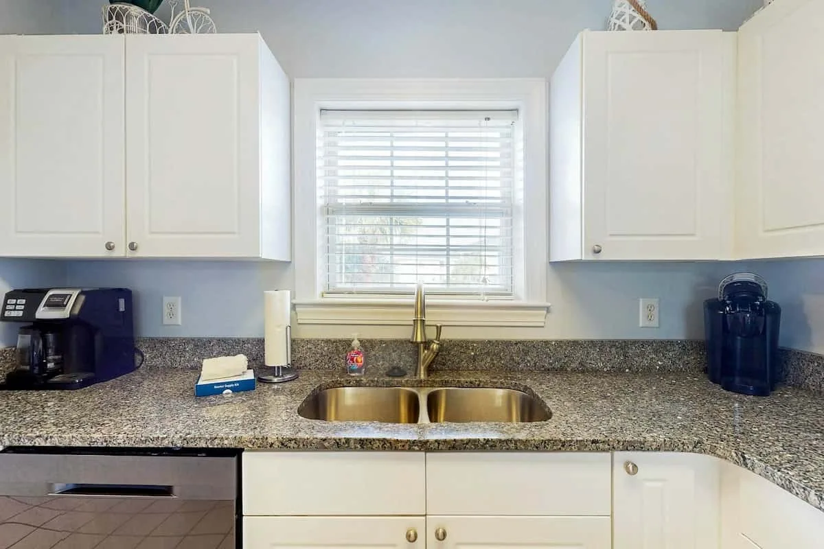 Una encimera de cocina con un fregadero está bajo una ventana, entre armarios blancos.