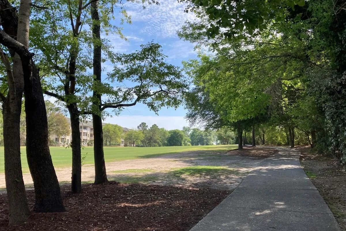 Un sendero pavimentado atraviesa un parque frondoso junto a un campo abierto.