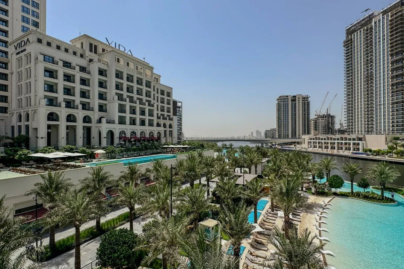 Una plaza urbana ajardinada con palmeras, piscinas y edificios modernos de gran altura.