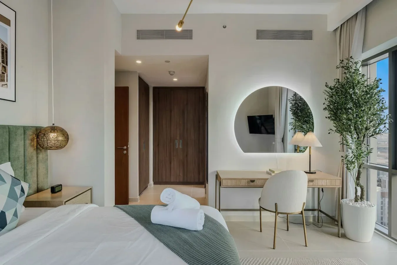 Un dormitorio luminoso con cama, escritorio y espejo redondo.