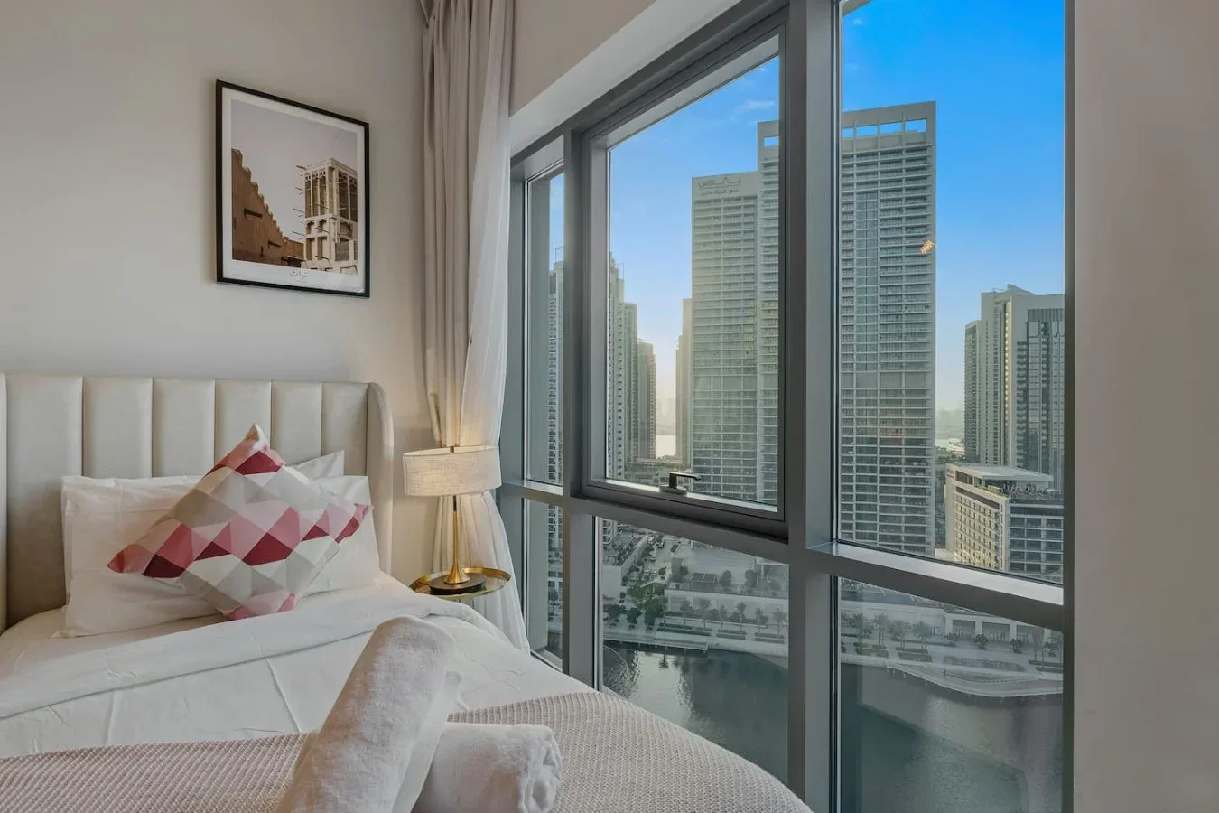 Dormitorio con una cama junto a grandes ventanas con vistas a edificios de la ciudad.