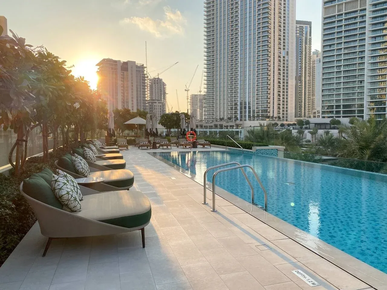 Una piscina al aire libre junto a una terraza pavimentada, asientos reclinables y edificios altos.