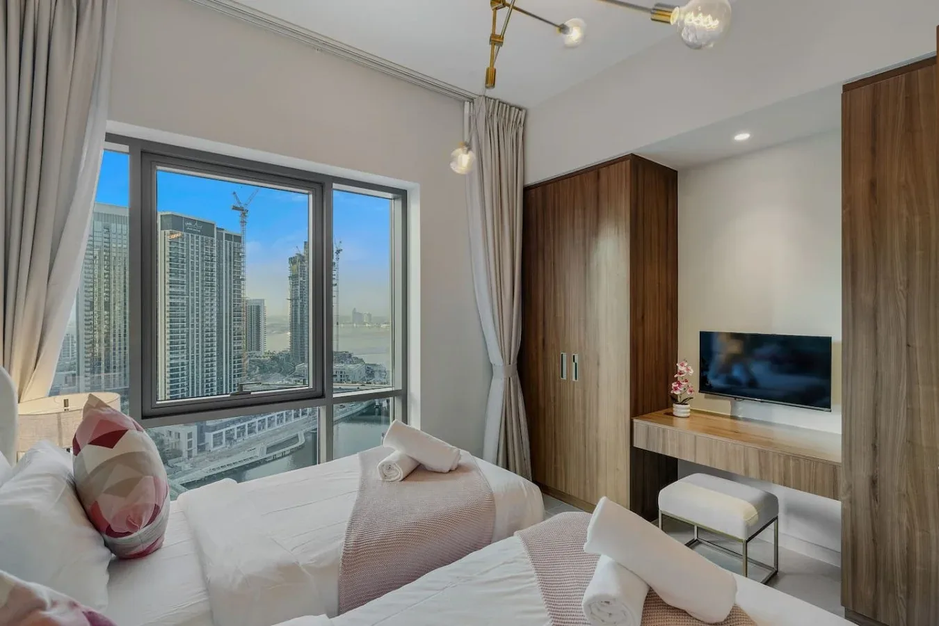 Un dormitorio luminoso con ropa de cama blanca, grandes ventanas y edificios altos a la vista.