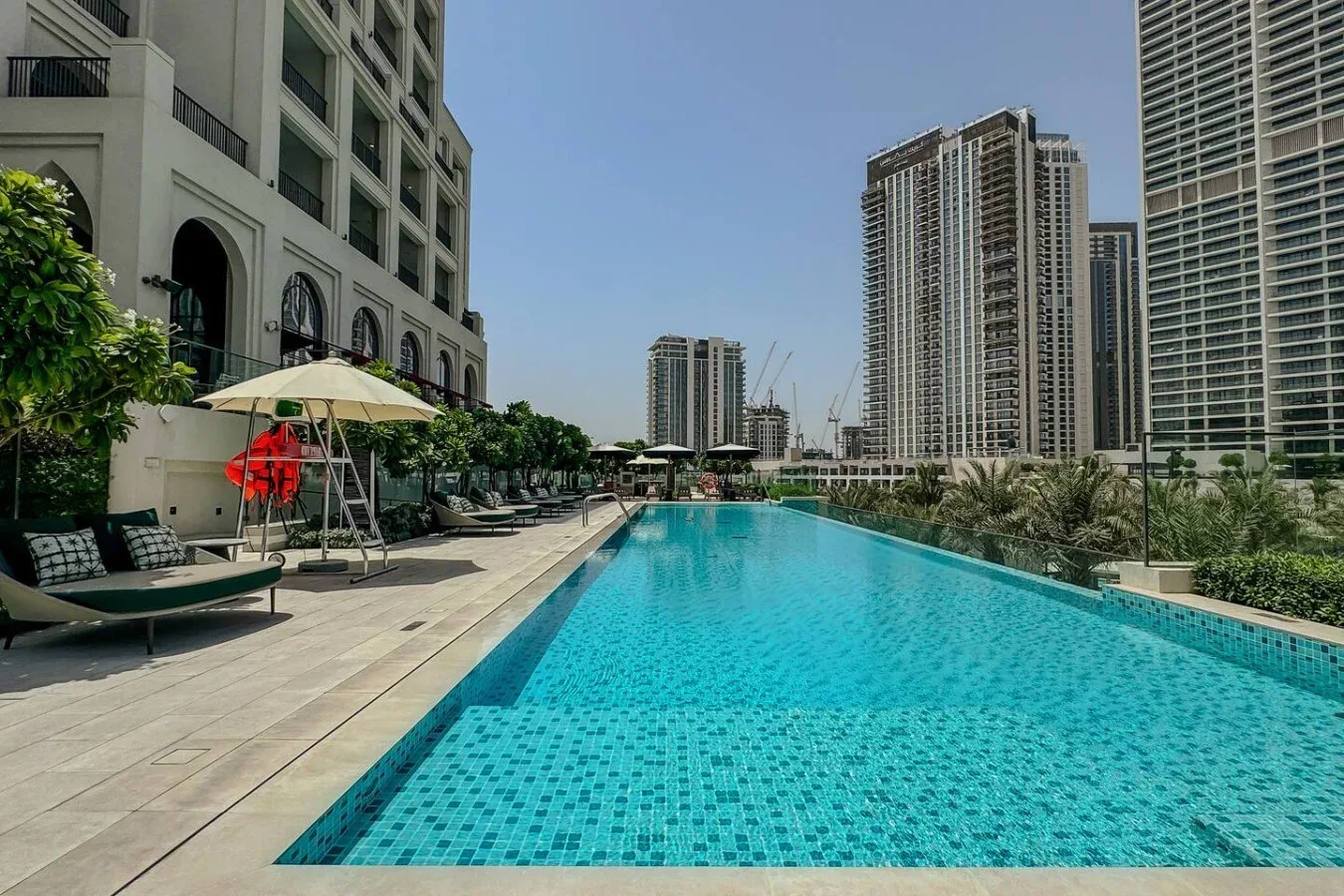 Una piscina al aire libre junto a edificios altos, con asientos de descanso en la terraza.