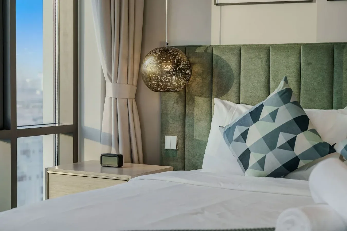 Dormitorio con cama, cojín decorativo geométrico y una ventana junto al cabecero.