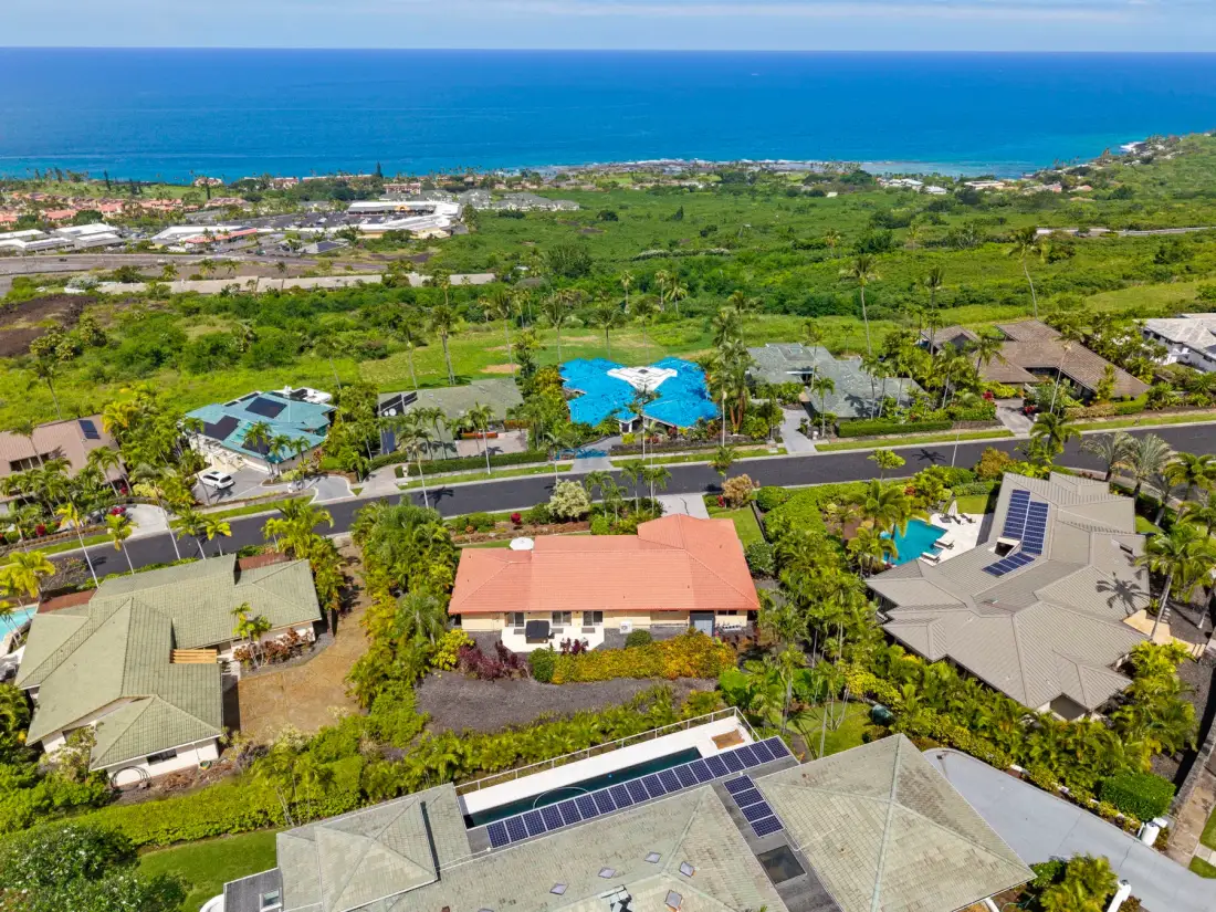 An aerial view of homes and pools among greenery, with the ocean in the distance.