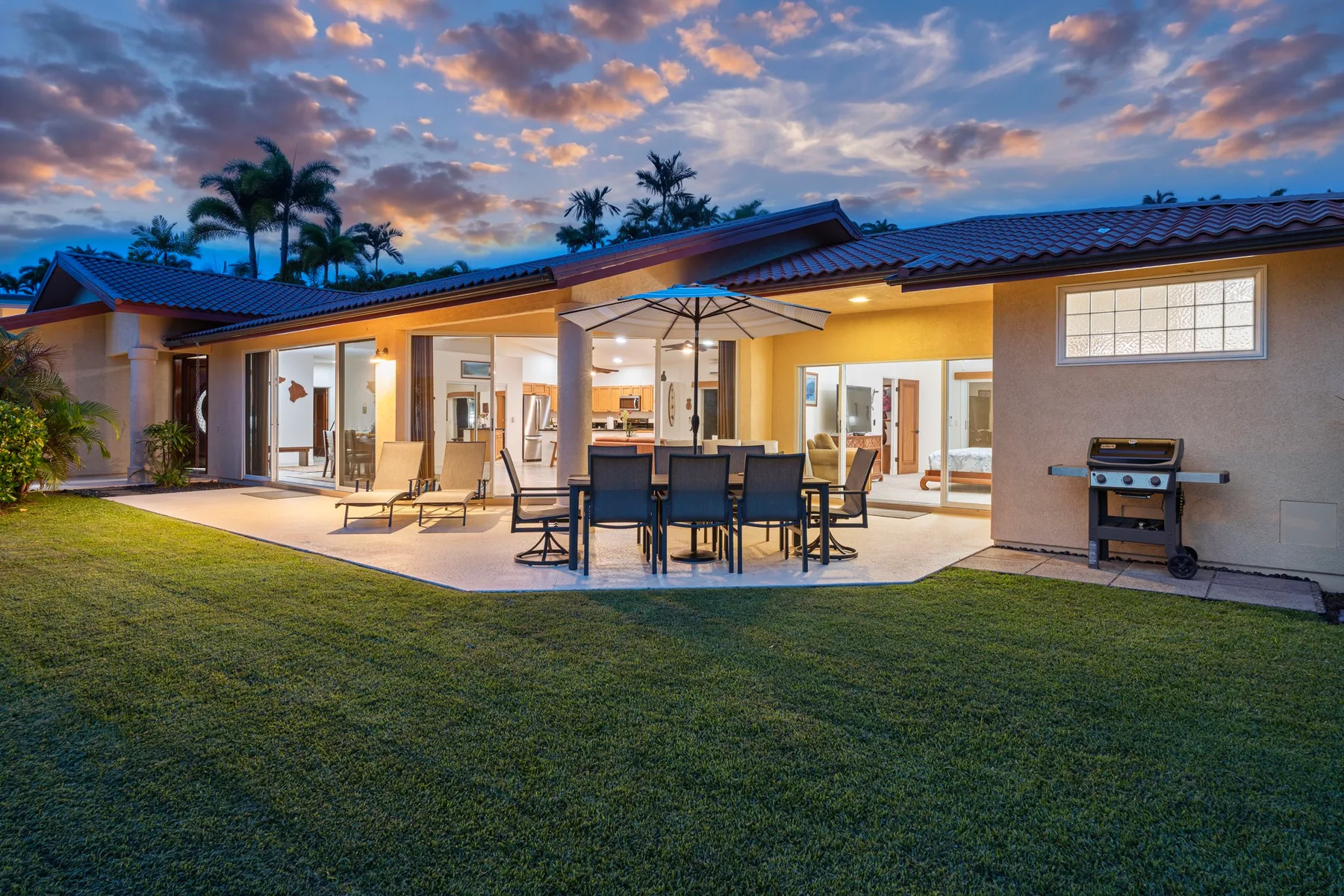 An illuminated patio with outdoor dining furniture, a grill, and a lawn at dusk.