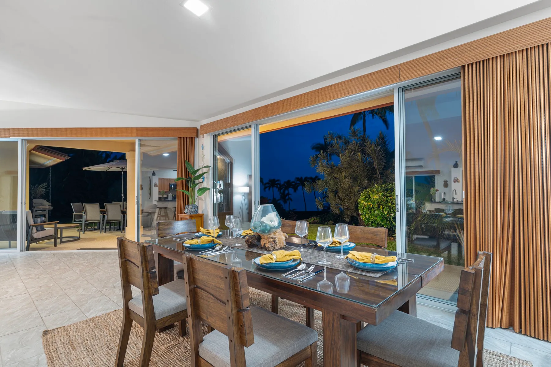A dining area with a wooden table beside wide windows overlooking palm trees.