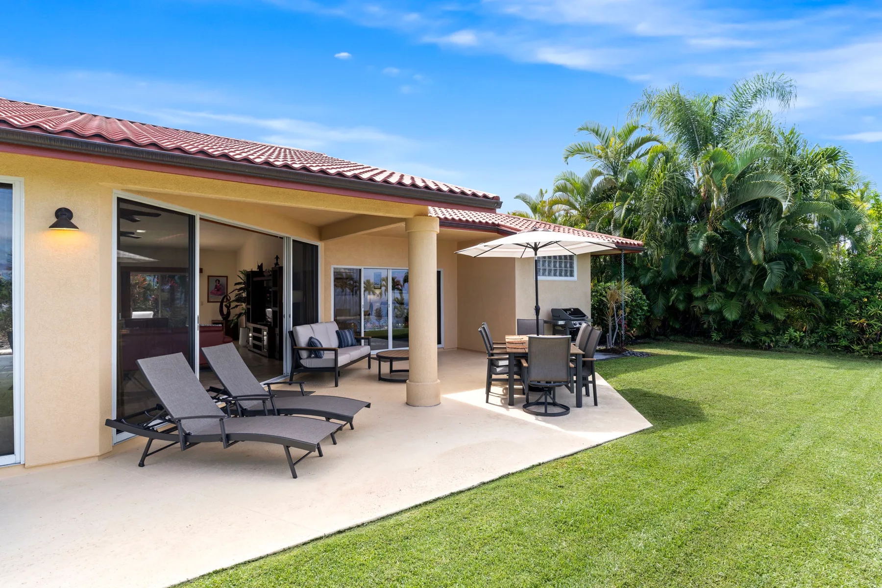 A covered patio with lounge seating and a dining table opens onto a green lawn.