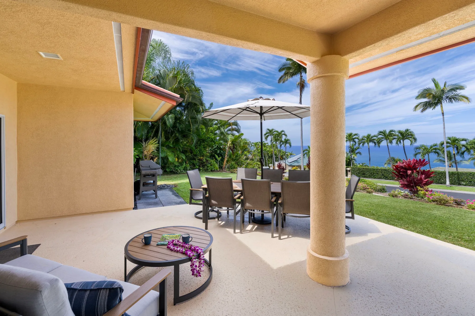 A covered outdoor patio with dining seating overlooks a landscaped garden and the ocean.