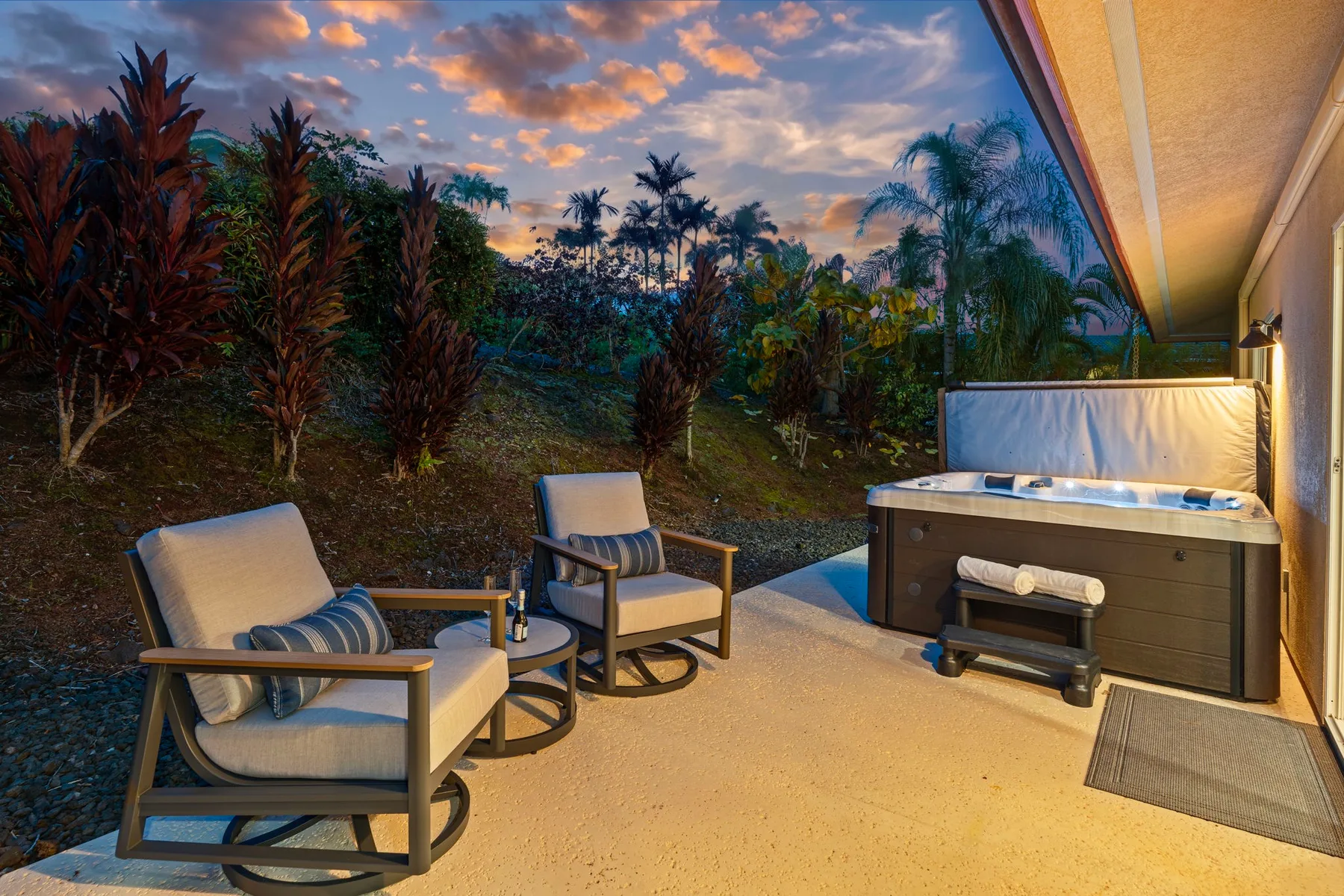 An outdoor patio has cushioned seating and a hot tub beside tropical plants.